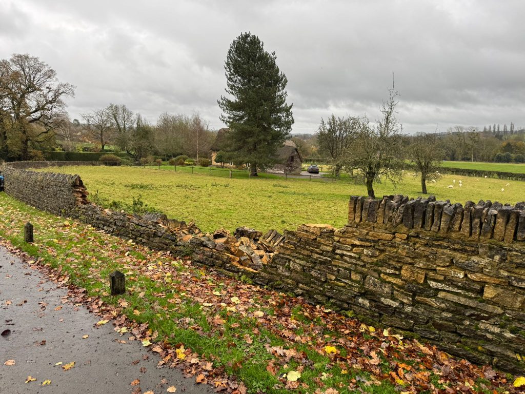Collapsed section of a Cotswold stone roadside wall, with fallen stones on the verge before JPJ rebuilt it