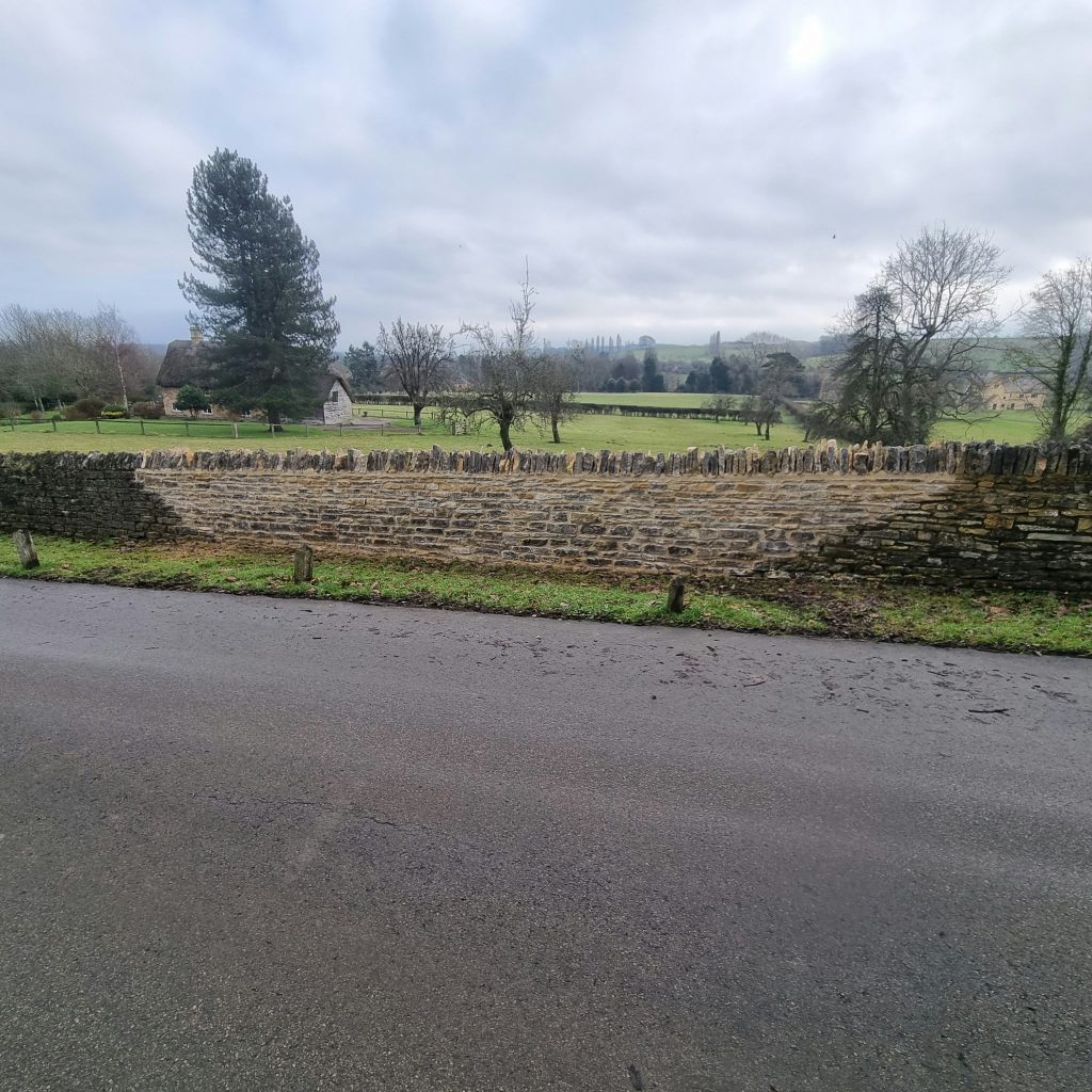 Newly rebuilt section of Cotswold stone roadside wall with a traditional upright-stone coping, joining the weathered original wall on either side